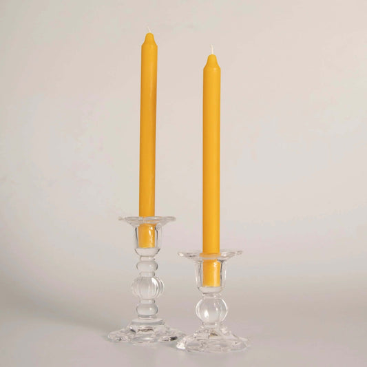 Deux bougie plus précisément "Deux cierges" en cire d'abeille jaunes, faits artisanalement à la main en Gaspésie, Québec par Florimel & Cie , placés dans des bougeoirs en verre transparent.